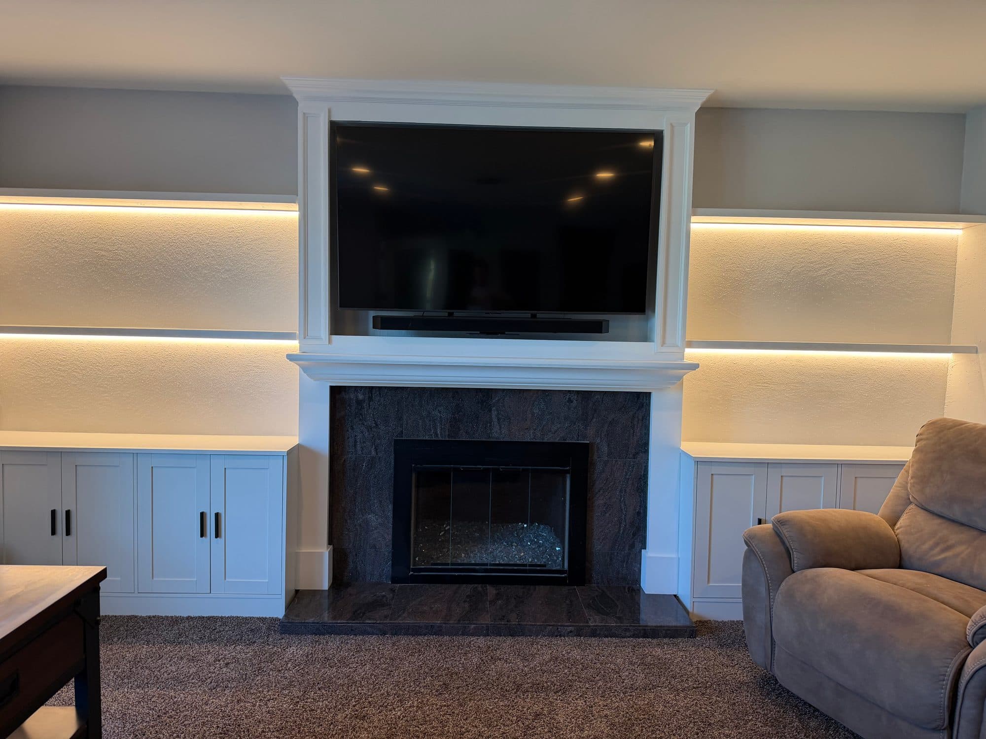 White fireplace media wall with TV in a trim surround, LED-lit floating shelves and white base cabinets on both sides of a dark tile fireplace