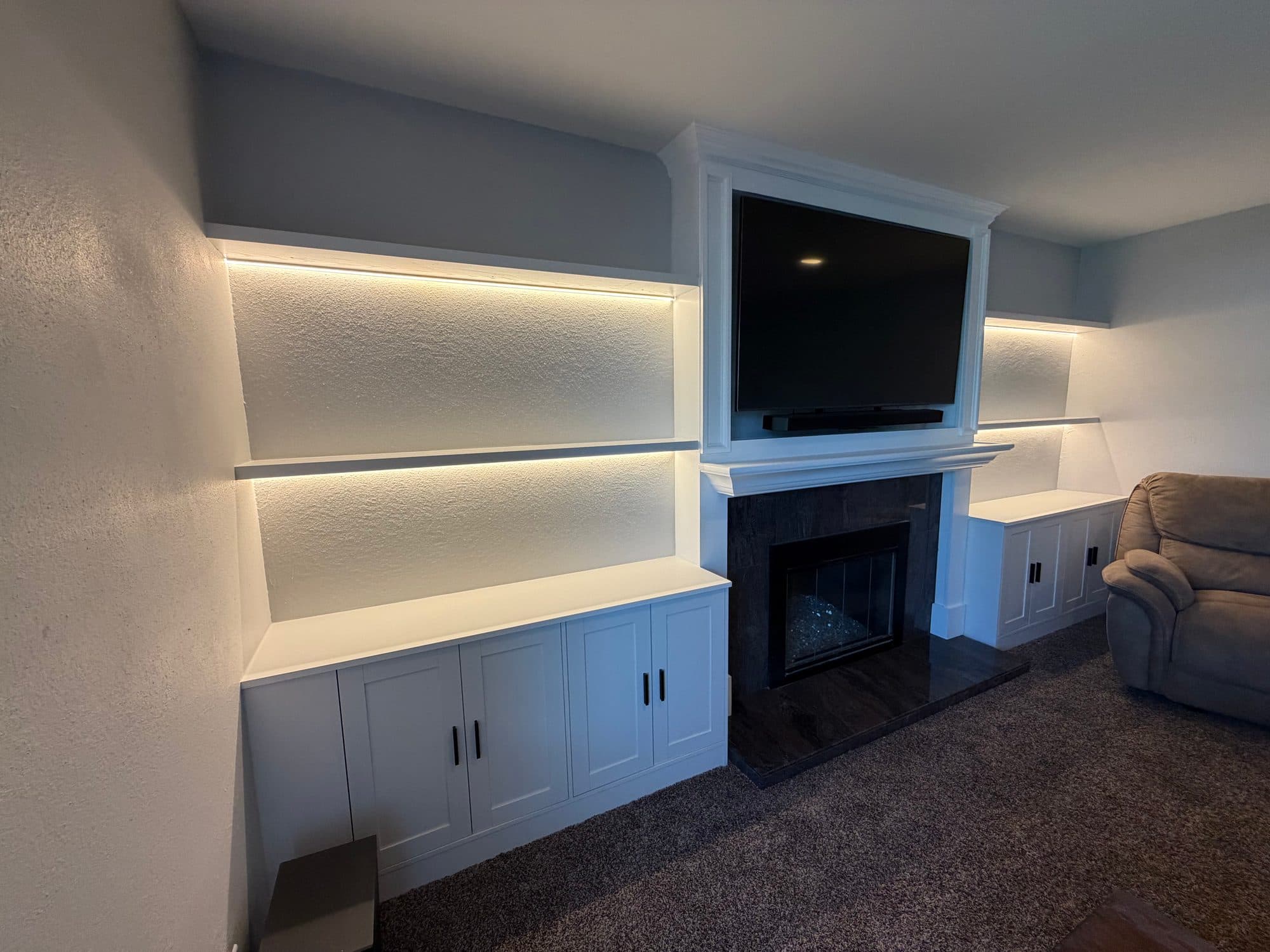 Angled view of a white built-in media wall with LED-lit recessed shelves, base cabinets and a framed TV above a tile fireplace