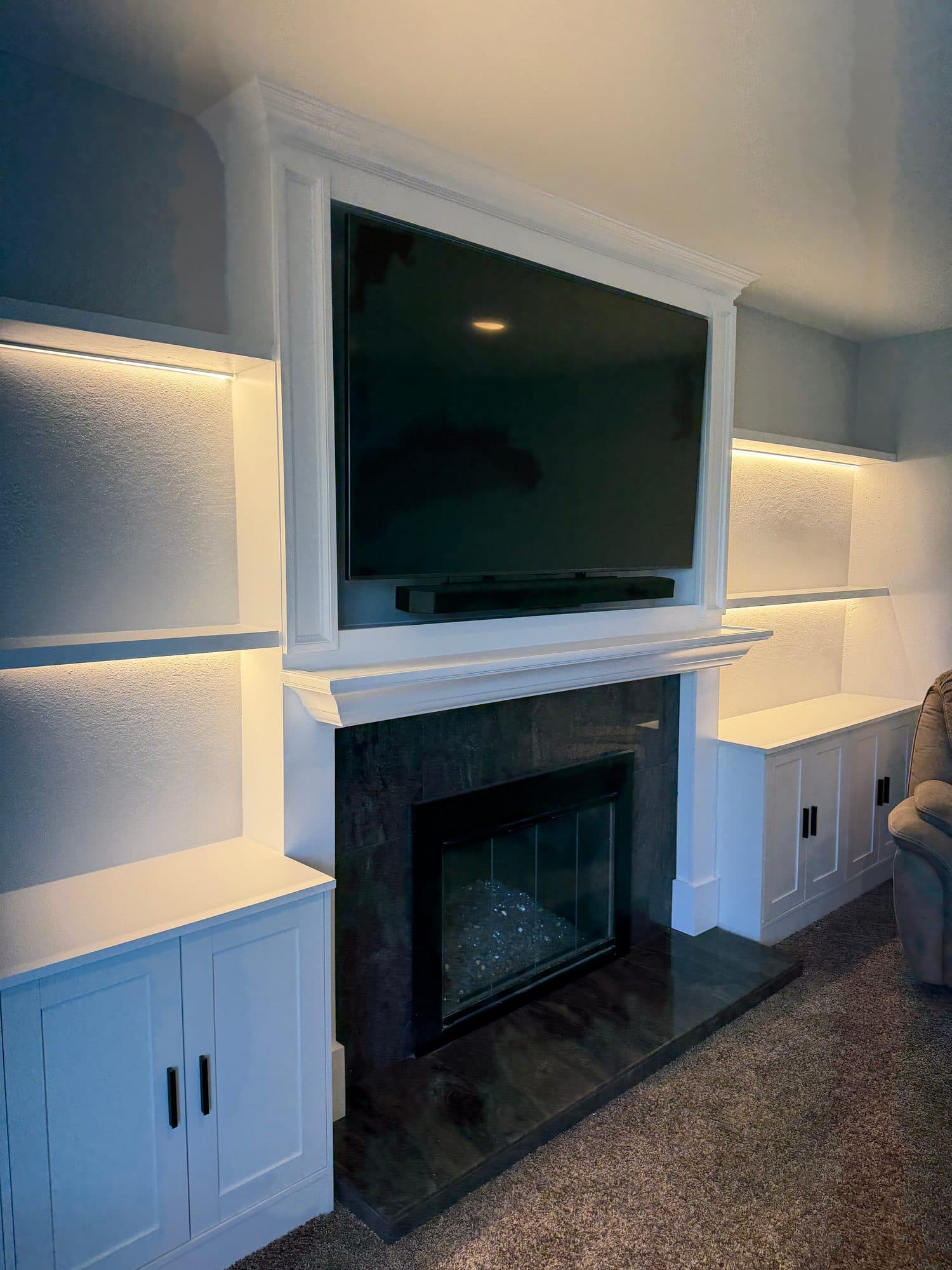 Angled view of a white fireplace media wall with crown molding, framed TV and lit side niches