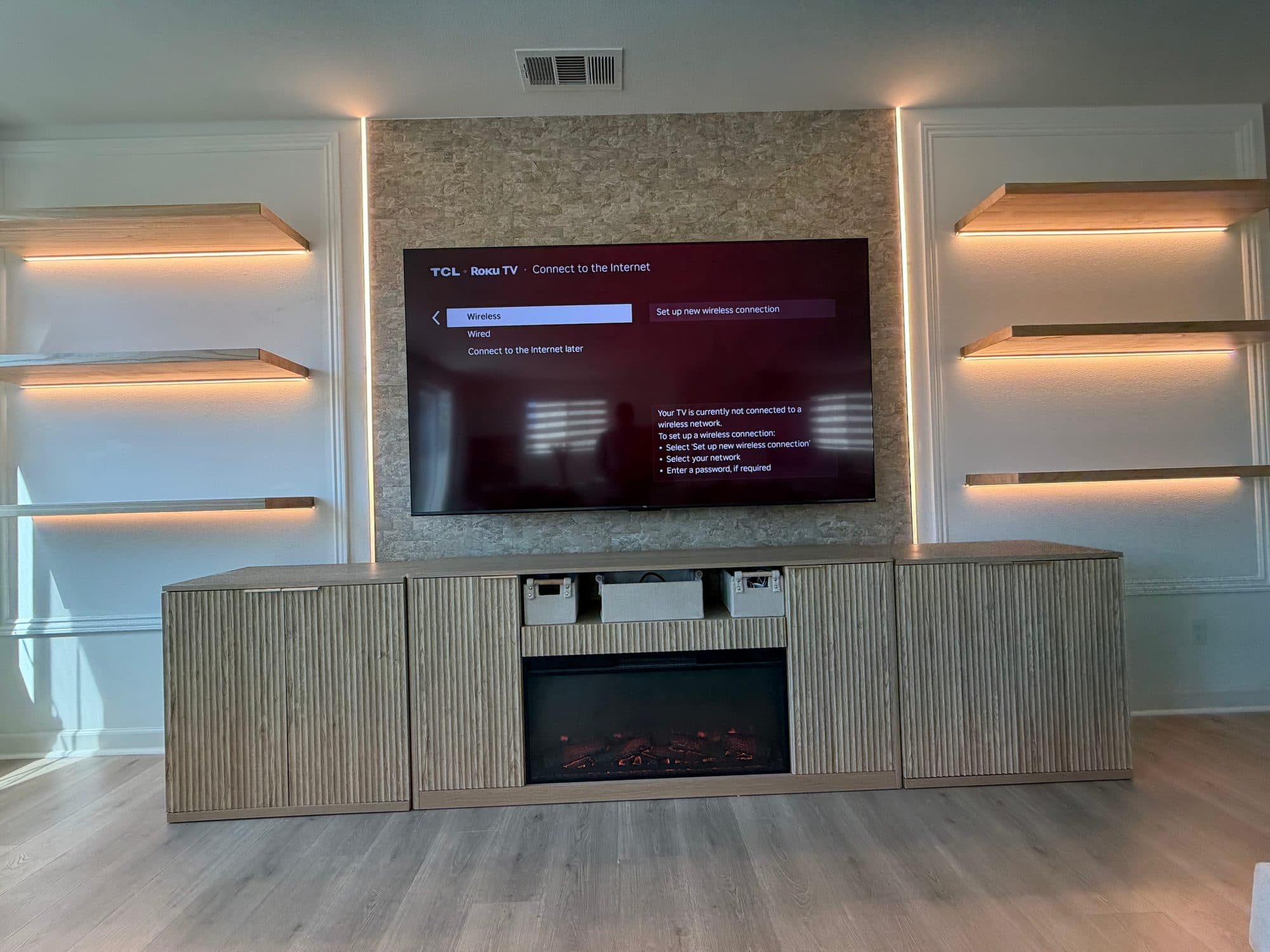 TV mounted on a stacked stone panel flanked by white panel walls with LED-lit oak floating shelves and a fluted wood console with electric fireplace