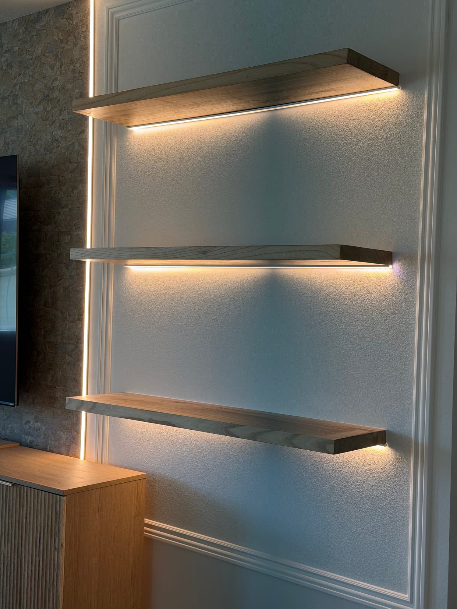 Three LED-lit oak floating shelves on a white wall with picture-frame molding beside a stone TV panel