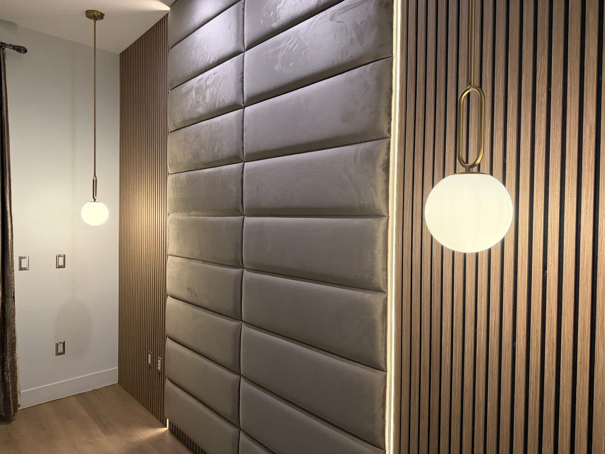 Angled view of gray upholstered wall panels next to a dark wood slat section with a globe pendant light